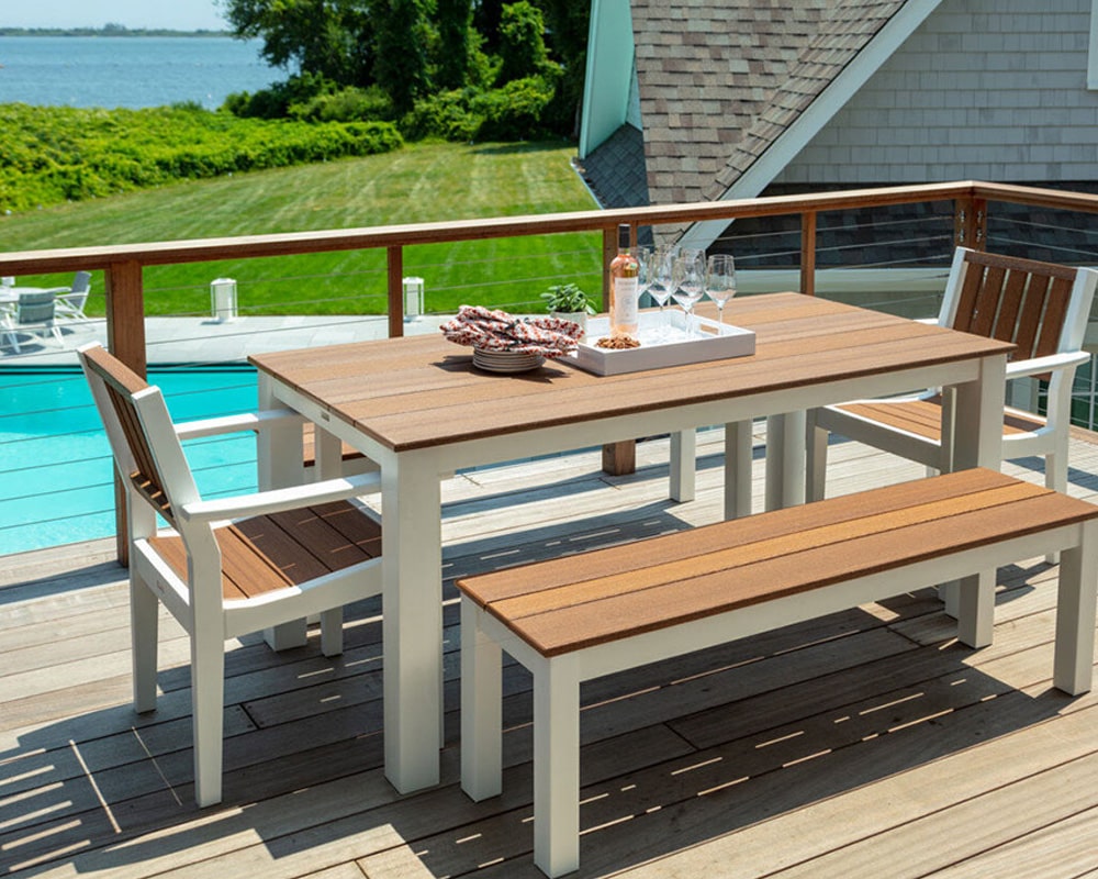 Modern outdoor dining set with a wooden table, chairs, and bench on a sunny patio with a pool and green landscape